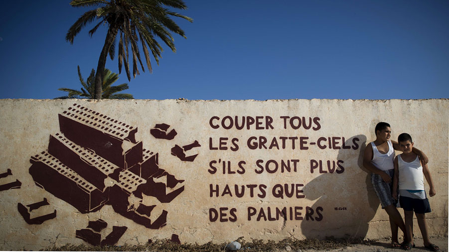 Children stand by a mural by Polish artist M-CITY decorating a wall in the village of Erriadh, on the Tunisian island of Djerba, on August 7, 2014, as part of the artistic project "Djerbahood". Artists from 34 diffrents nationalities were invited by France-based Tunisian artist Mehdi Ben Cheikh to take part in an initiative to turn Djerba's Erriadh district into an "open sky museum". Mural reads "Cut all the skyscrapers if they are taller than palm trees".  AFP PHOTO / JOEL SAGET = RESTRICTED TO EDITORIAL USE, MANDATORY MENTION OF THE ARTIST UPON PUBLICATION, TO ILLUSTRATE THE EVENT AS SPECIFIED IN THE CAPTION =